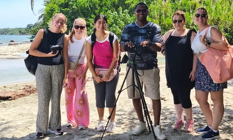 Six smiling travelers on a beach with their guide and a spotting scope on a tripod